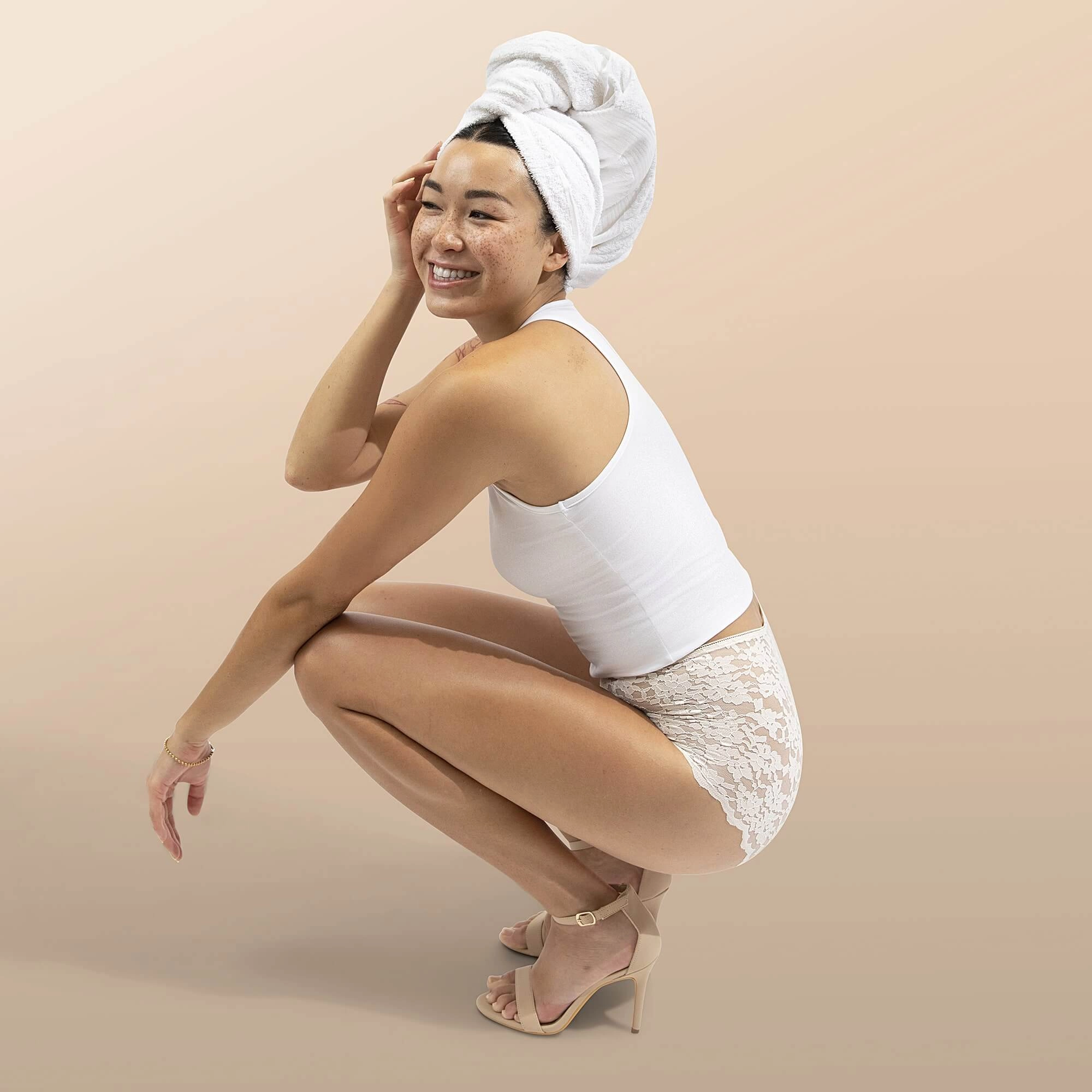 Woman in a white tank and cream lace high-waisted briefs, crouching and smiling with a towel wrapped on her head